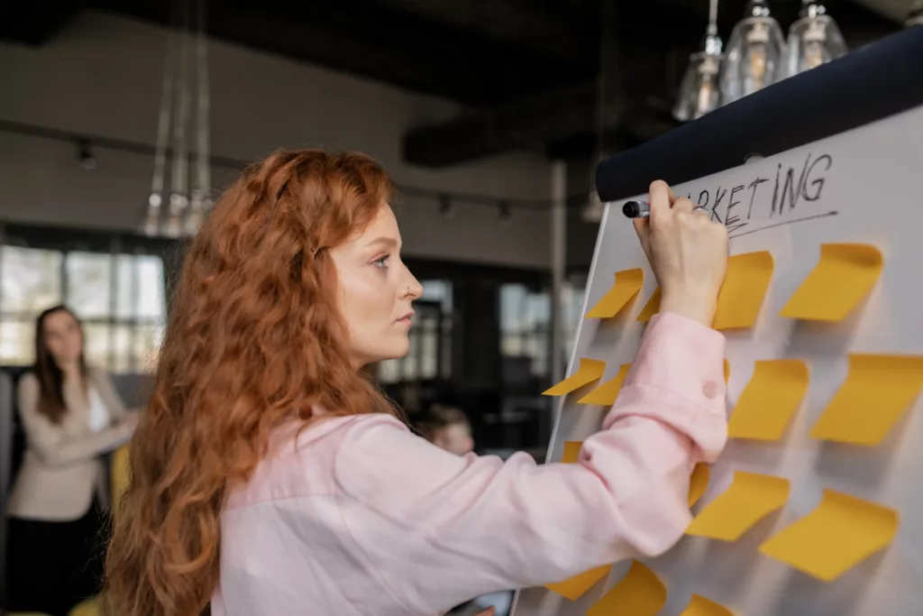 a woman in front of a drawing board that has marketing written on it with sticky notes all over the board.